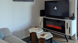 Een woonkamer met een flatscreen-tv, een open haard, een houder voor een afstandsbediening en een klein tafeltje met een boek.
