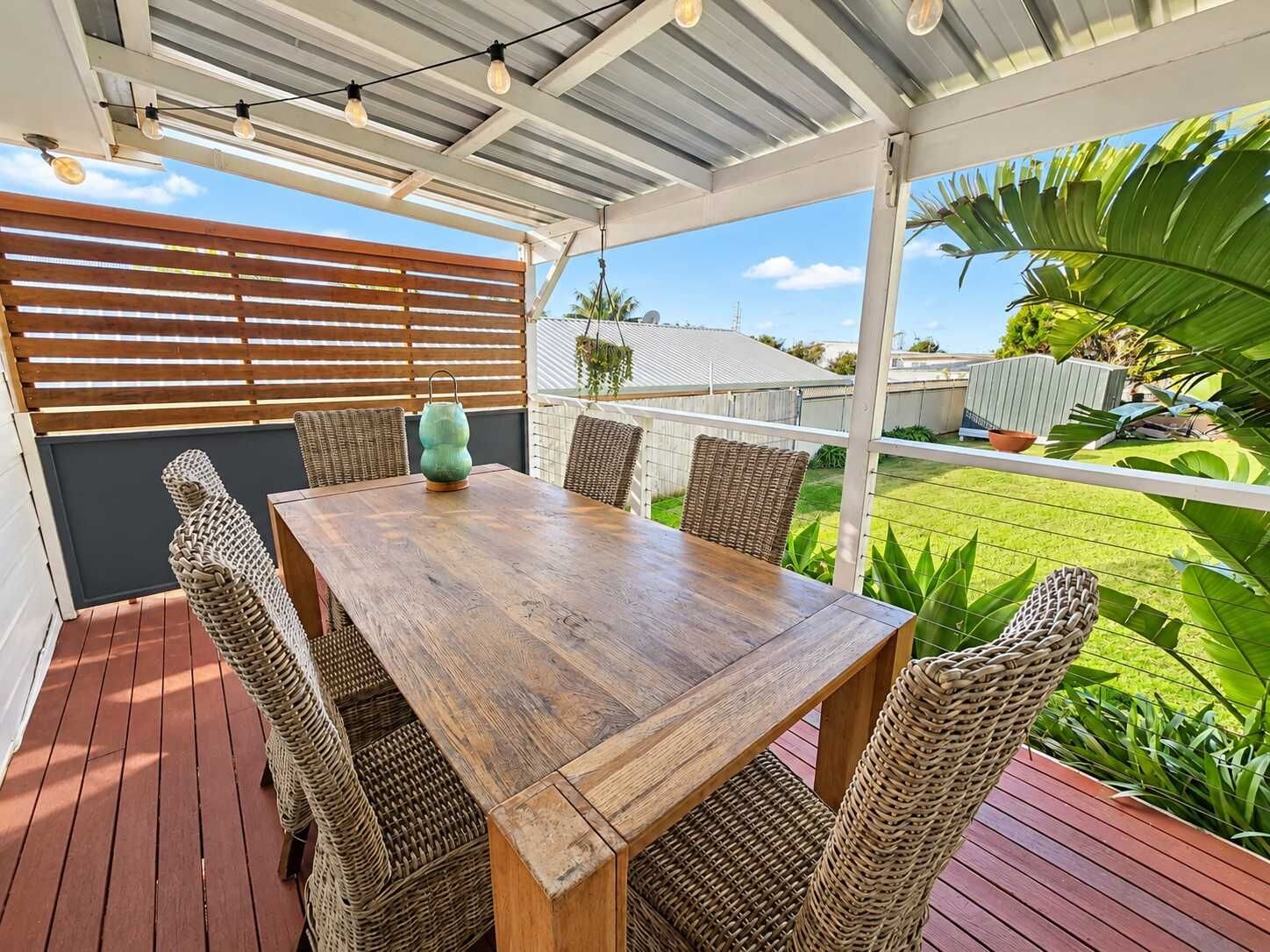 An outdoor patio with a wooden table and wicker chairs, a hanging light fixture, and a view of a green lawn and house.