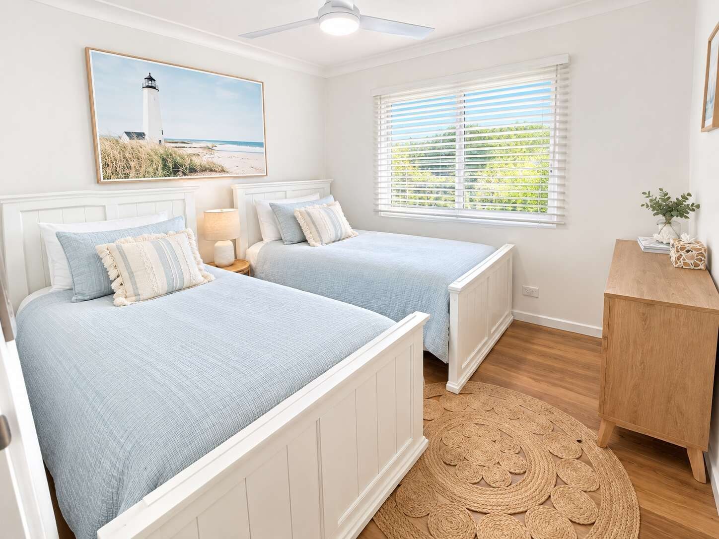 A bedroom with two beds, a ceiling fan, a window with blinds, a bedside table with a lamp, and a framed picture on the wall.