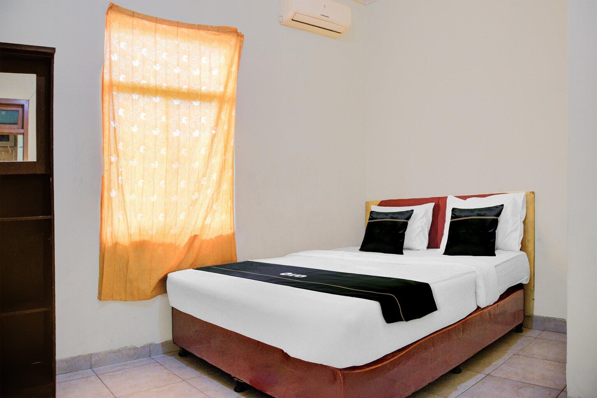 Una cama individual con estructura de madera, colchón blanco y almohadas negras y rojas. Se observa una unidad de aire acondicionado pequeña en la pared sobre la cama.