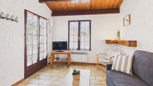 Een woonkamer met een houten plafond, een grijze bank, een televisie op een houten dressoir en een plant in een pot op een tafel.