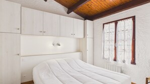 Een slaapkamer met een bed, houten balken in het plafond, witte muren en een raam met gordijnen.