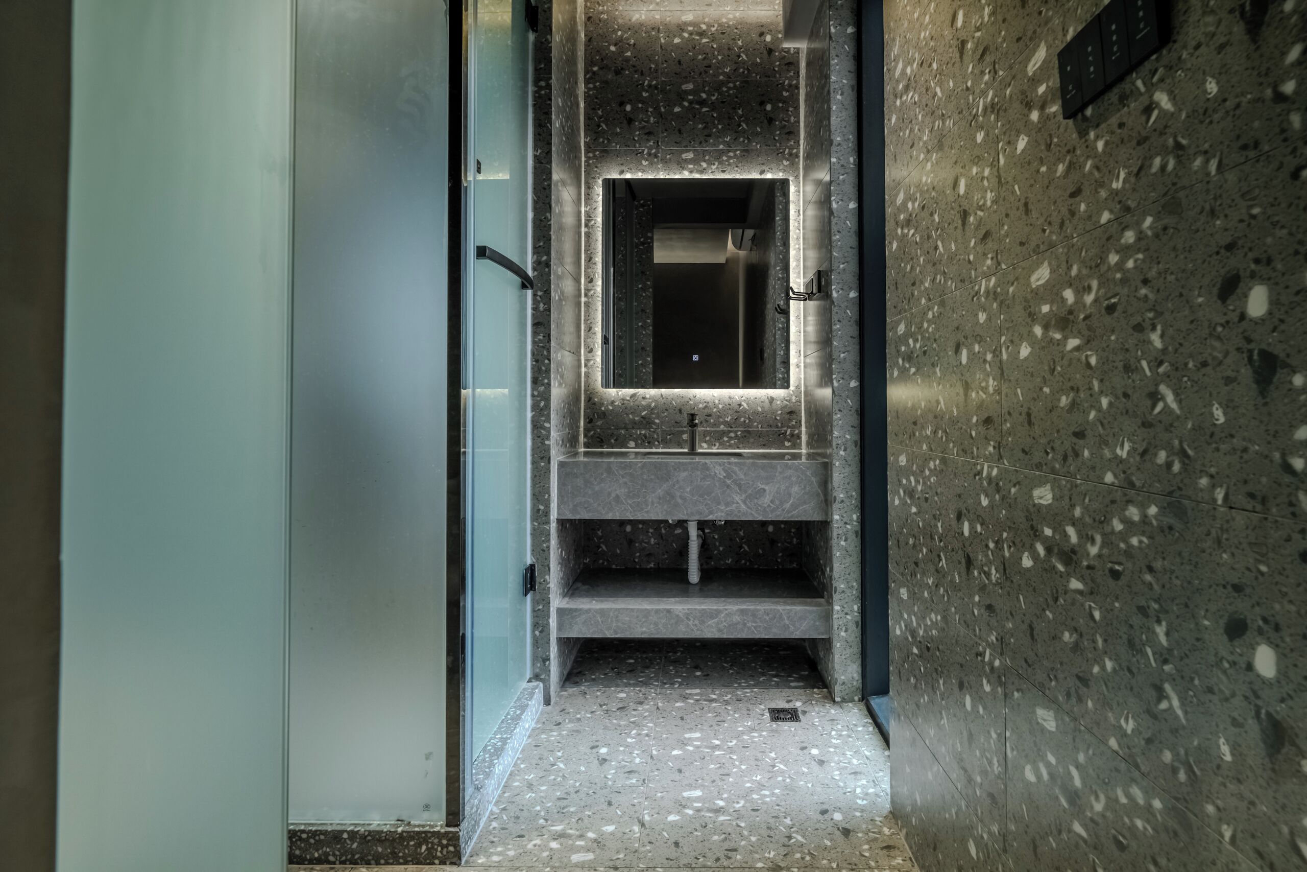 A narrow hallway with a modern bathroom featuring a glass door, a marble sink, and a speckled floor.