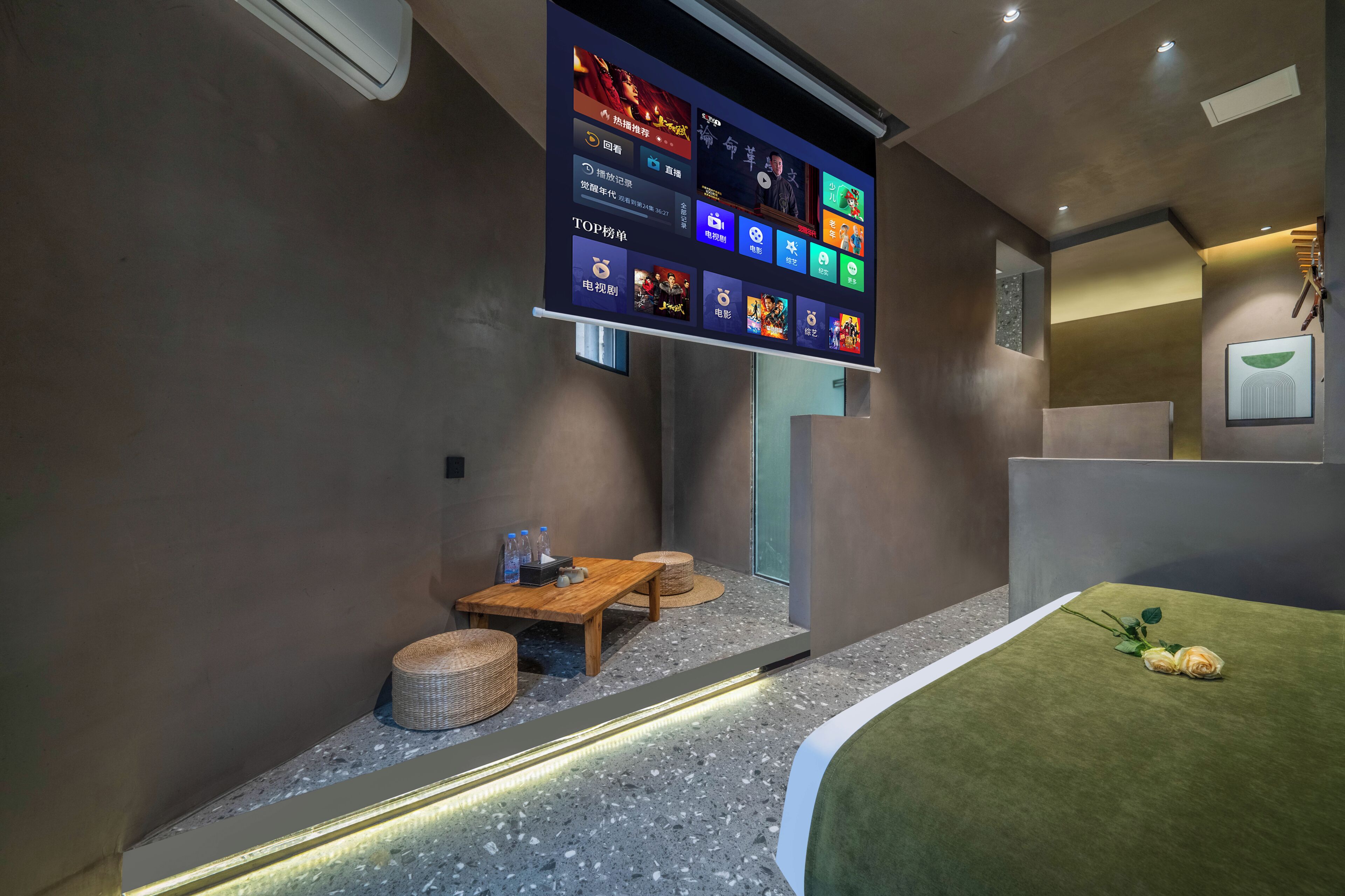 Habitación de hotel moderna con una gran pantalla digital mostrando diverso contenido, una cama con cubierta verde, una pequeña mesa de madera con una cesta y un aire acondicionado instalado en la pared.