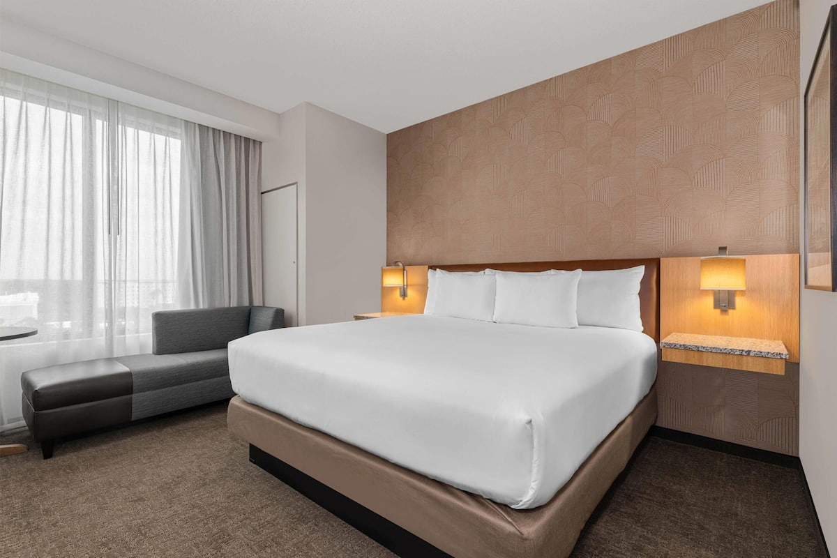 Quarto de hotel com uma cama grande, um sofá, duas luminárias de cabeceira, uma janela com cortinas e uma parede estampada.