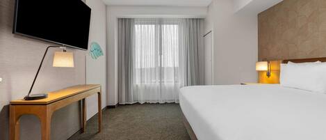 A hotel room with a bed, a desk with a lamp, a TV mounted on the wall, and a window with sheer curtains.
