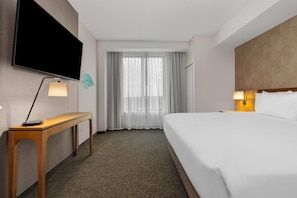 Quarto de hotel com uma cama, uma escrivaninha com abajur, uma TV na parede e uma janela com cortinas leves.