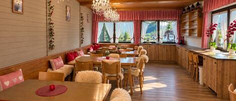 A cozy dining area with wooden tables and chairs, a chandelier, and large windows with curtains.