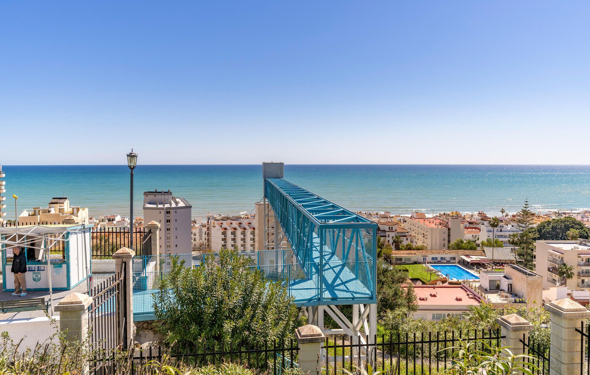 Eine Küstenstadtlandschaft mit einer markanten, blauen Brücke, Wohnhäusern und freiem Blick auf das Meer.