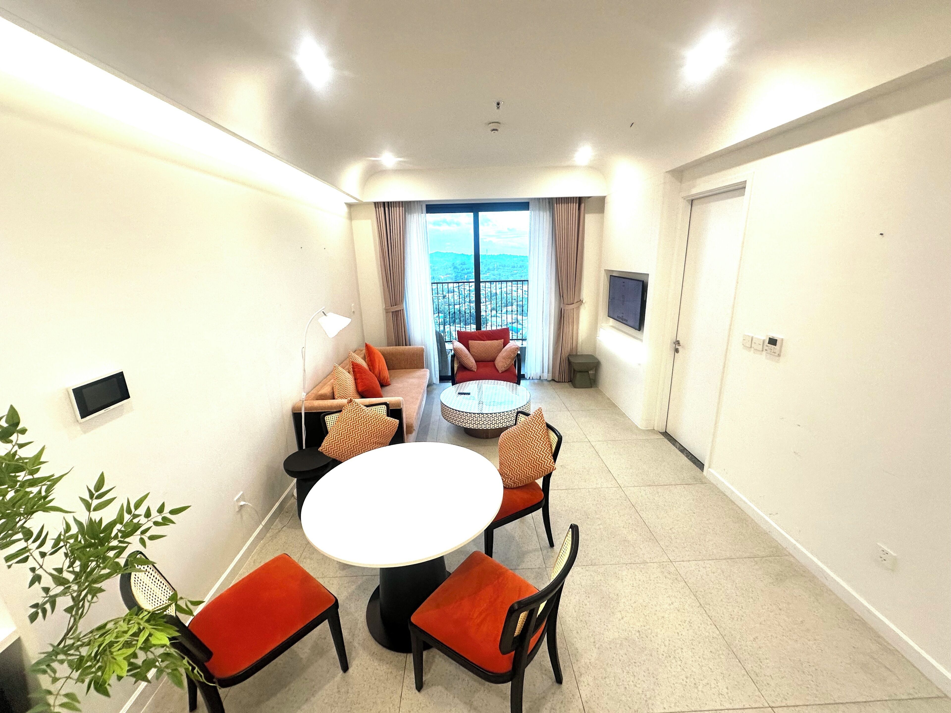 A modern hotel room with a balcony, a round dining table with chairs, a sofa, and a TV mounted on the wall.