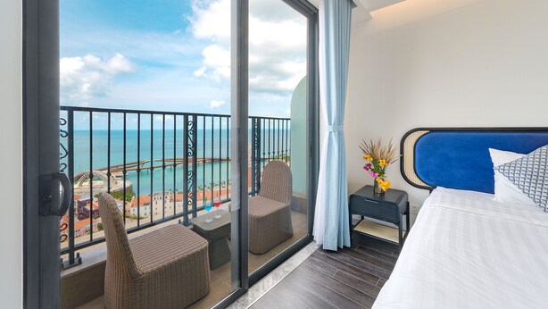 A hotel room with a balcony, a bed, a small table with a vase of flowers, and a view of the sea.