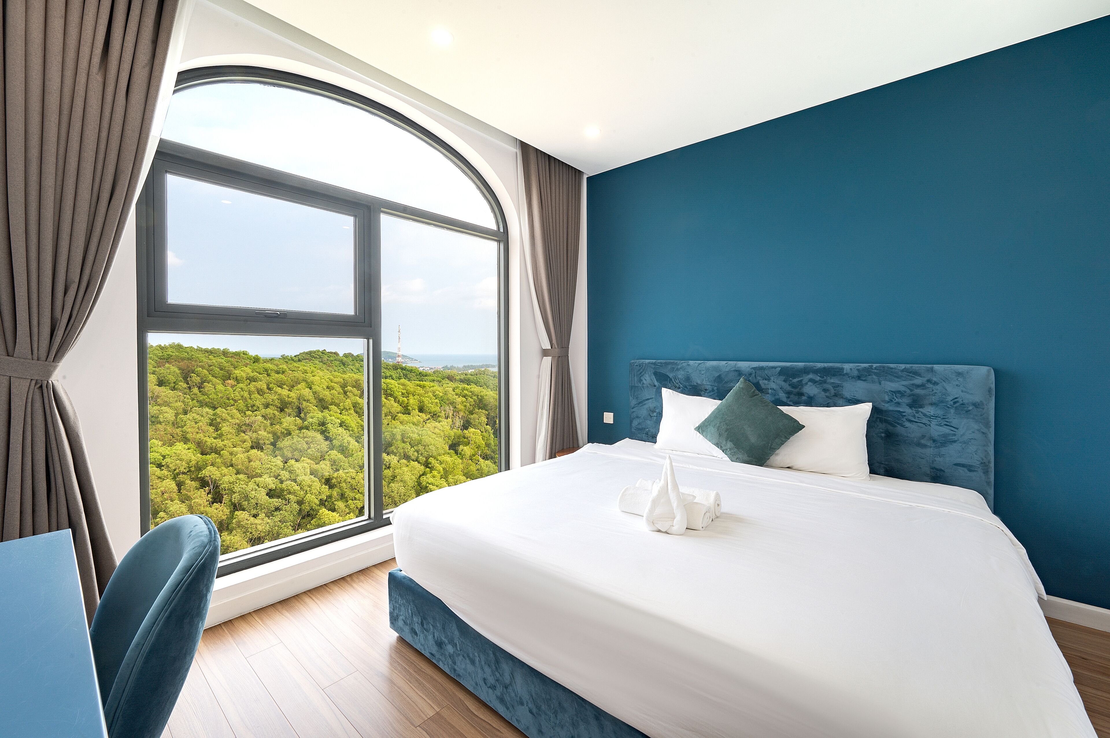 A hotel room with a large window, a bed with white linens, and a blue headboard.