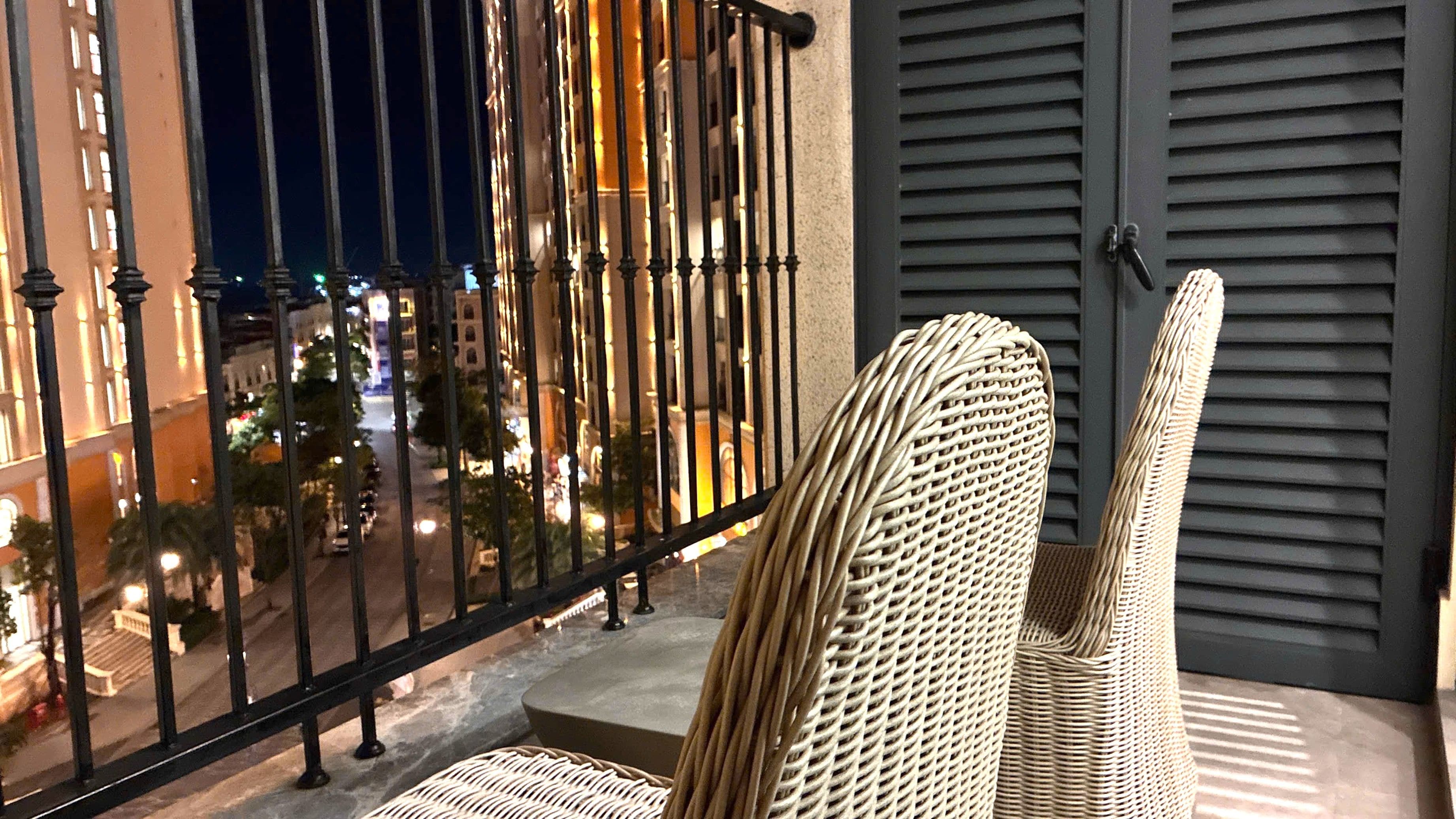 A balcony with a wicker chair, a view of a cityscape at night, and a building with grey shutters.