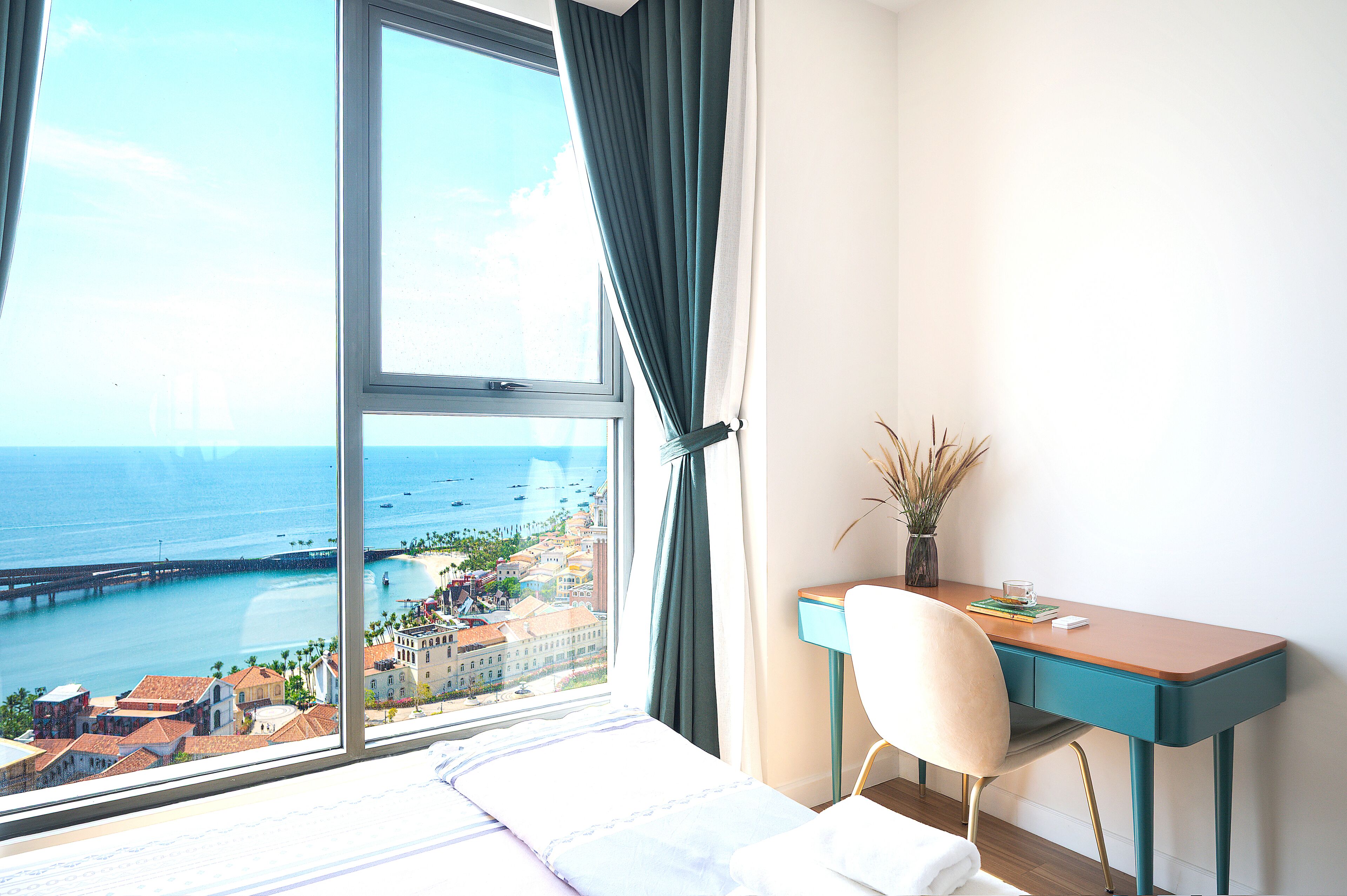 Une chambre d’hôtel avec une grande fenêtre, un bureau, une chaise et une vue sur la mer et les bâtiments.