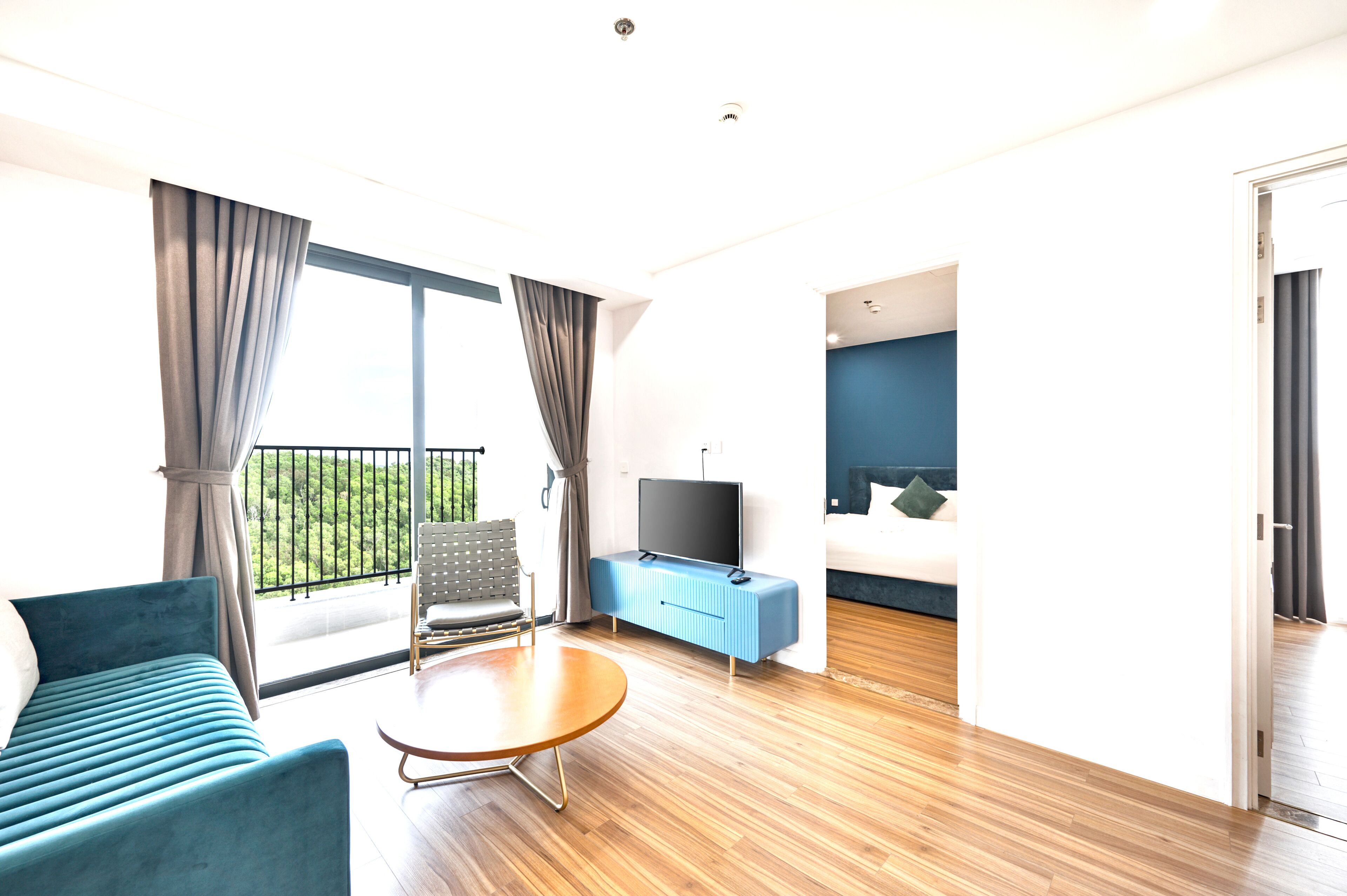 Habitación de hotel moderna con sofá, televisor sobre un mueble, mesa de comedor y balcón con vistas.