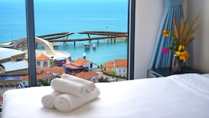 A hotel room with a bed, a window with a view of the sea and a bridge, and a vase with flowers on a nightstand.