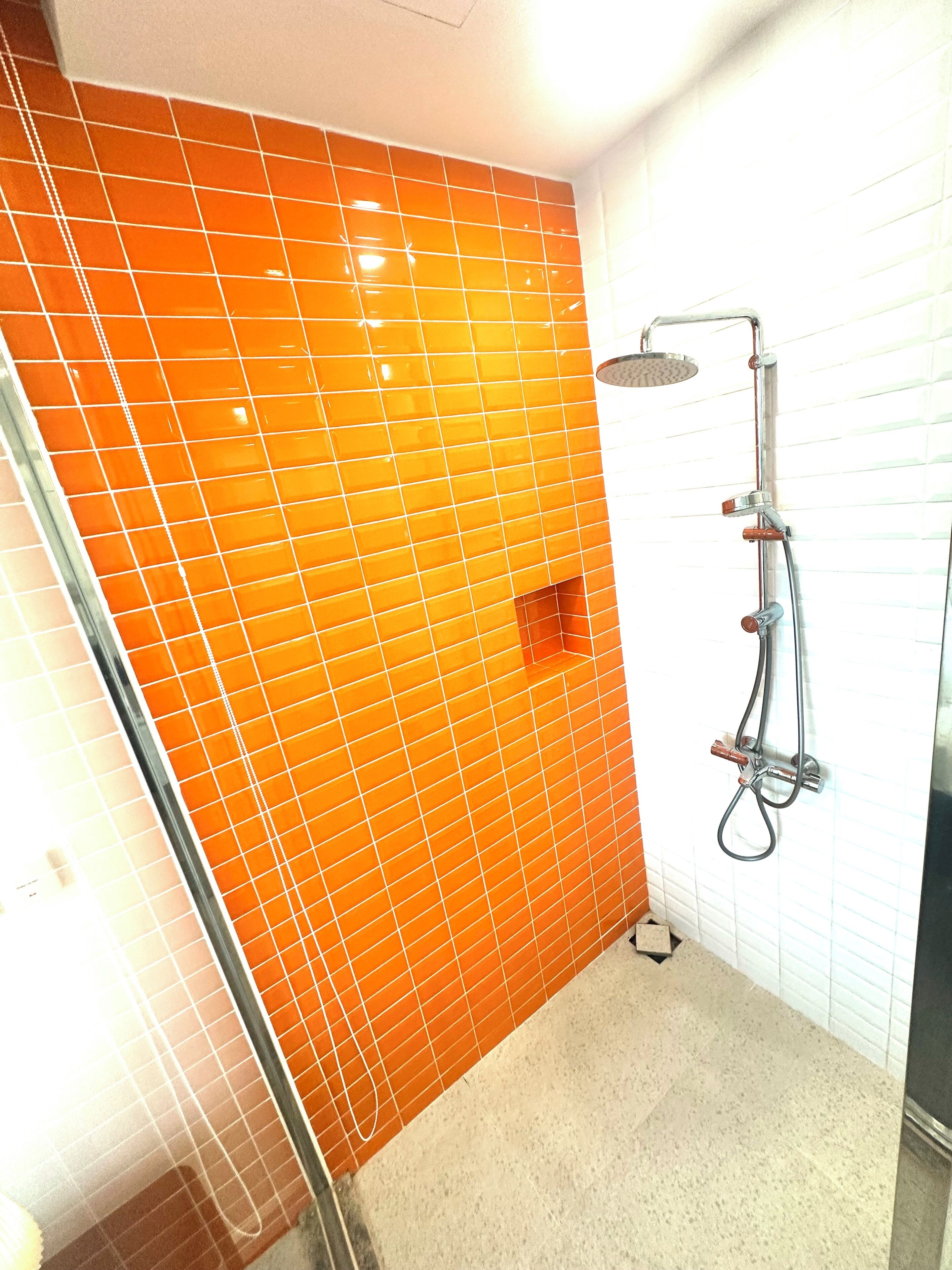 A bathroom with orange tiled walls, a shower with a handheld showerhead, and a glass shower door.