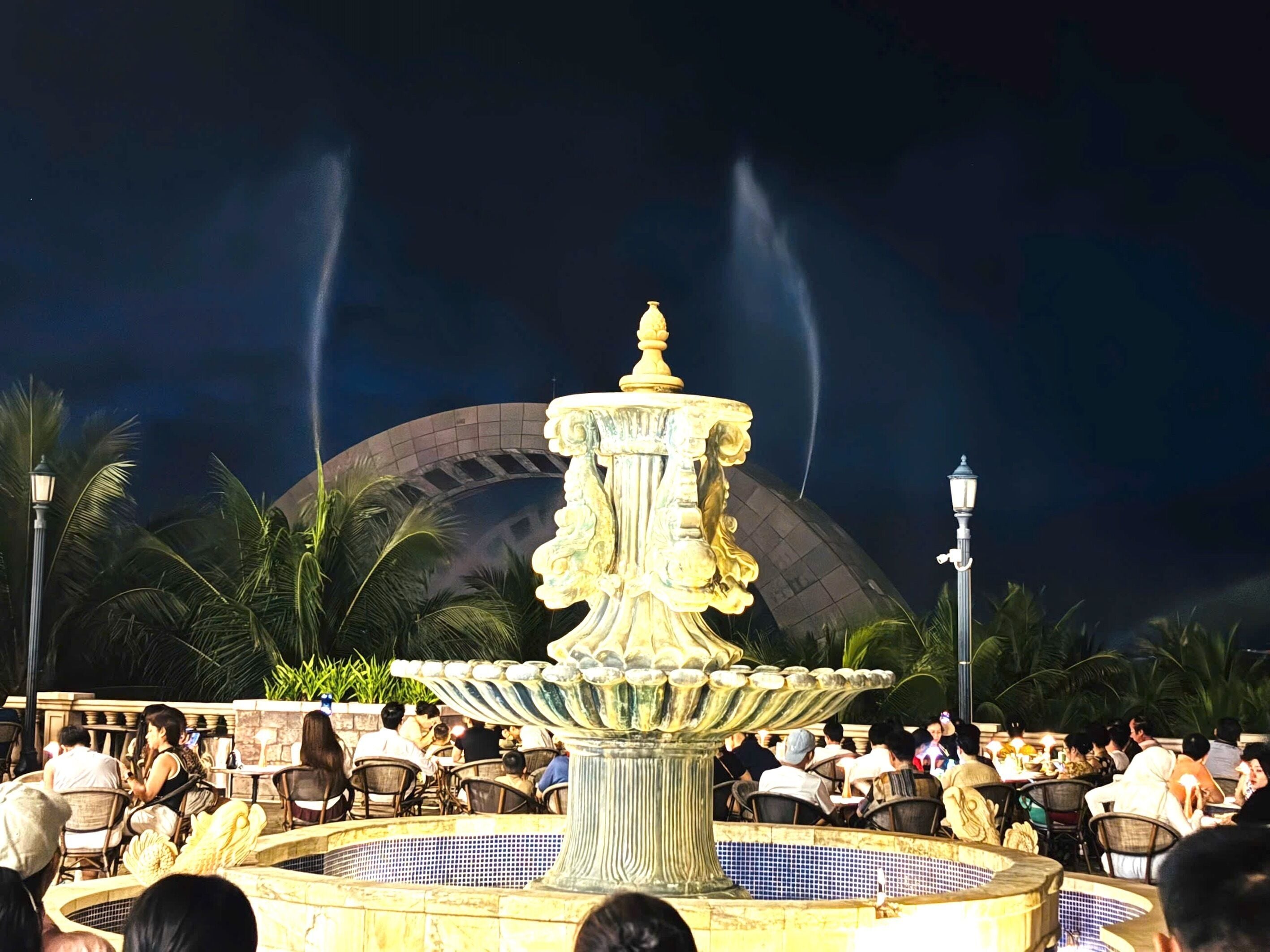 Une fontaine à plusieurs niveaux, entourée de palmiers et de personnes dînant à des tables.