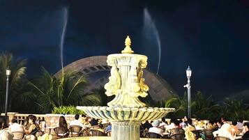 Une fontaine à plusieurs niveaux, entourée de palmiers et de personnes dînant à des tables.