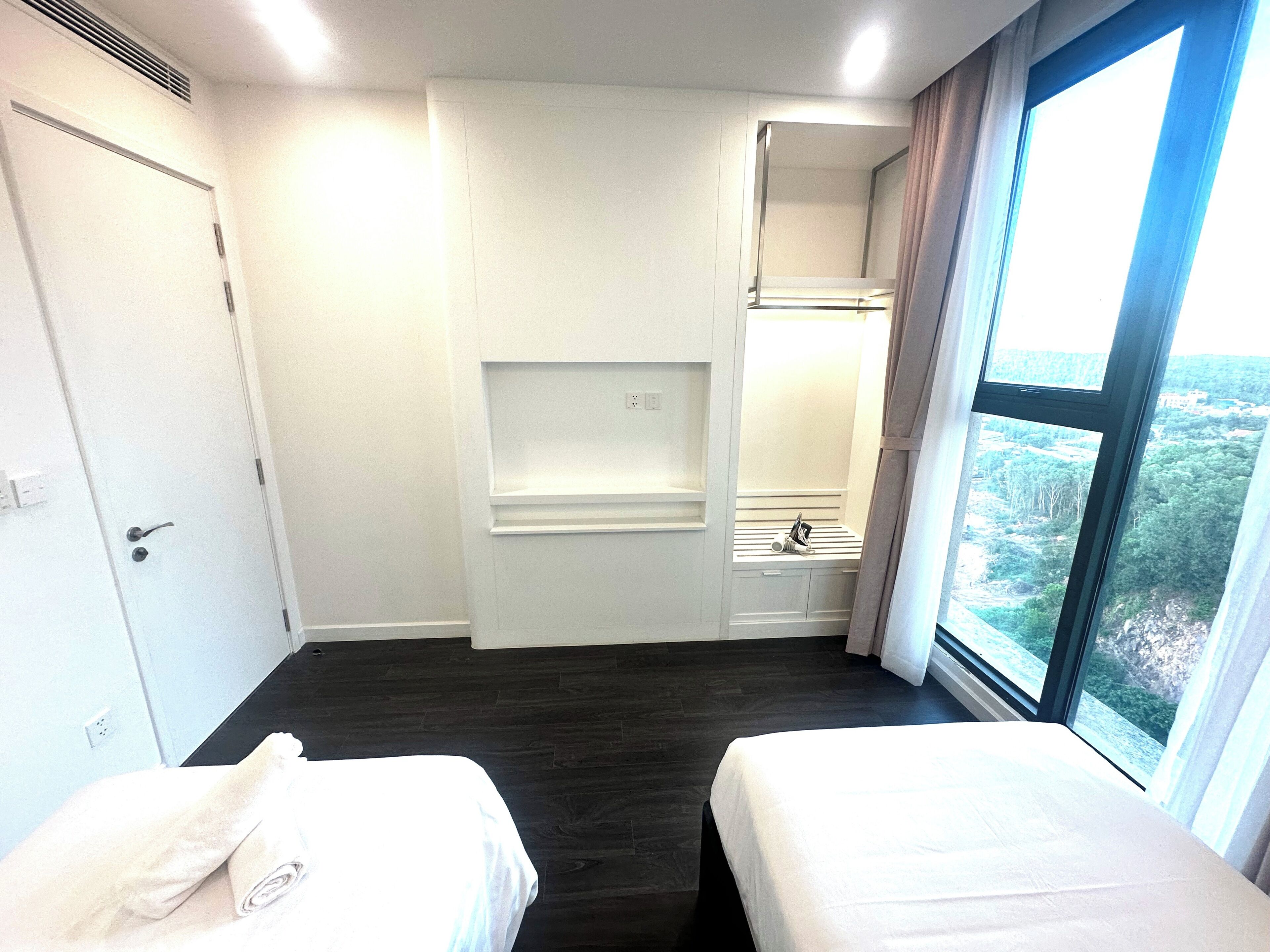 A hotel room with two beds, a built-in wardrobe, a small bathroom, and a large window offering a view.