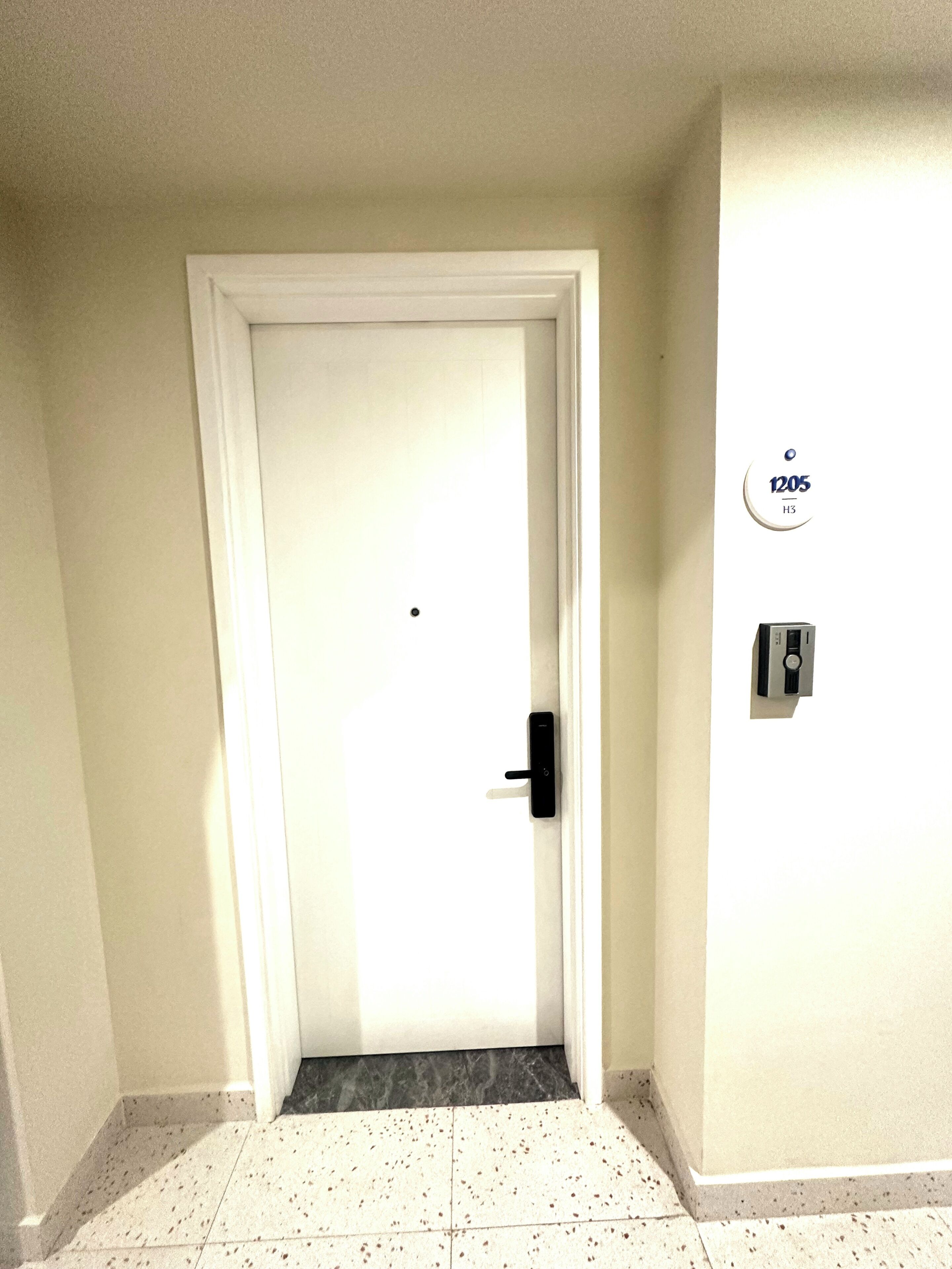A hotel room entrance with a white door, a small step leading to the floor, a wall-mounted intercom, and a sign with '1205 H3' on the wall.