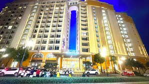 A multi-story hotel with a central vertical feature, a landscaped area with a water feature, and a group of people gathered in front.