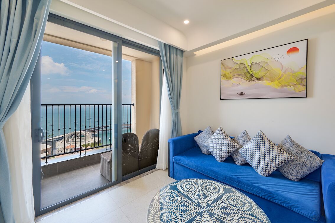A room with a blue sofa, patterned cushions, a large painting, and a view of the sea through a balcony.