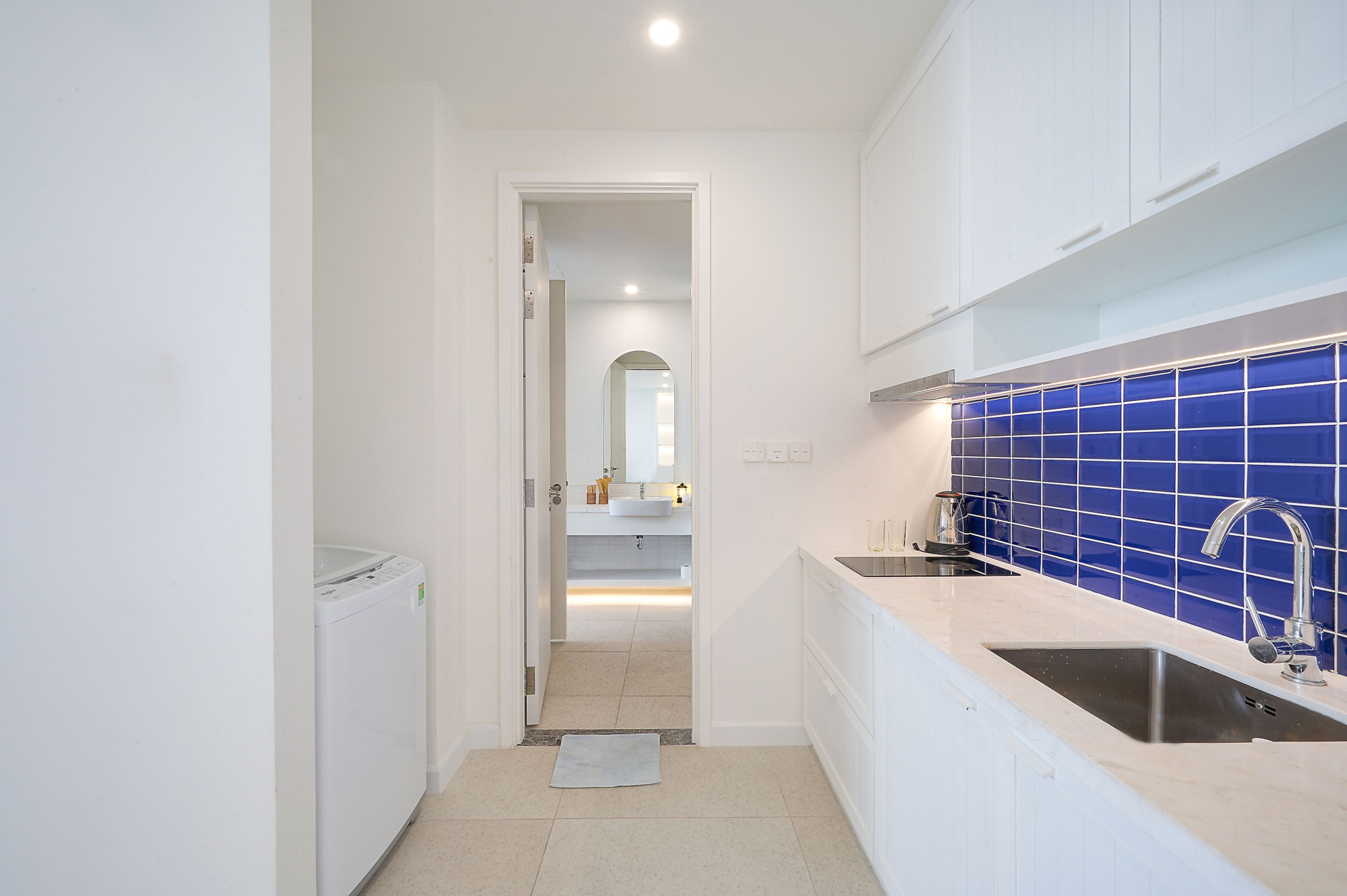 Un couloir étroit mène à une salle de bain où se trouvent une machine à laver blanche, un lavabo avec un pare-éclaboussures bleu et un miroir.
