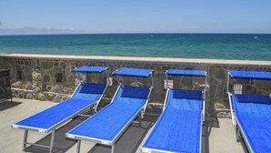 Une rangée de chaises longues bleues à armature métallique, alignées contre un mur de pierre, offre une vue sur une mer calme et des montagnes lointaines.