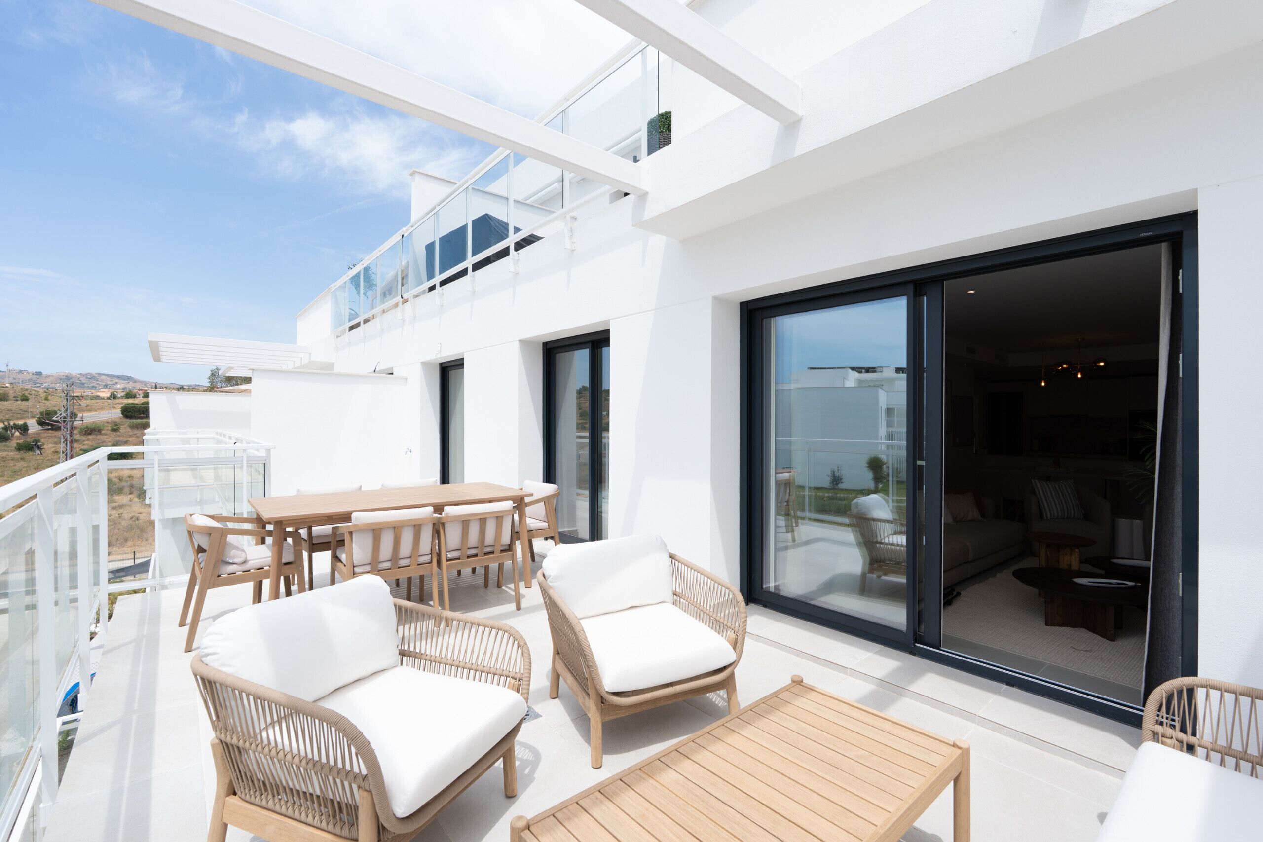Un patio exterior moderno con mobiliario de mimbre, una mesa de madera y una puerta corredera de cristal que da a un espacio interior.