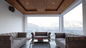 Habitación con techo de madera, ventanas amplias, dos sofás, una mesa de centro de vidrio y vistas a un paisaje montañoso.