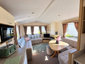 Interior compacto de uma motorhome, com sofá, televisão, cozinha compacta e área de refeições.