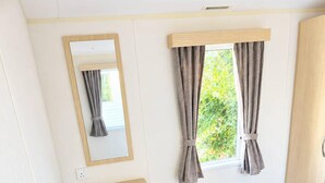 Un dormitorio con cama, ventana con cortinas, espejo, taburete pequeño y estante.