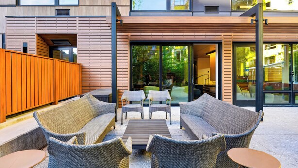 Un espace extérieur aménagé avec des meubles en osier et une table en bois, situé à côté d’un bâtiment doté de grandes portes vitrées.
