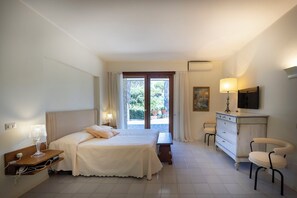 Ein Hotelzimmer mit Bett, Nachttisch, Lampe, Kommode, Stuhl und Blick ins Freie.