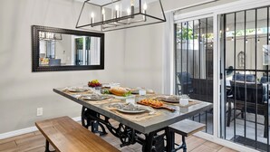 Uma sala de jantar com uma mesa grande posta para seis pessoas, um banco, um candelabro, um espelho e uma porta de vidro deslizante que dá para um pátio externo.