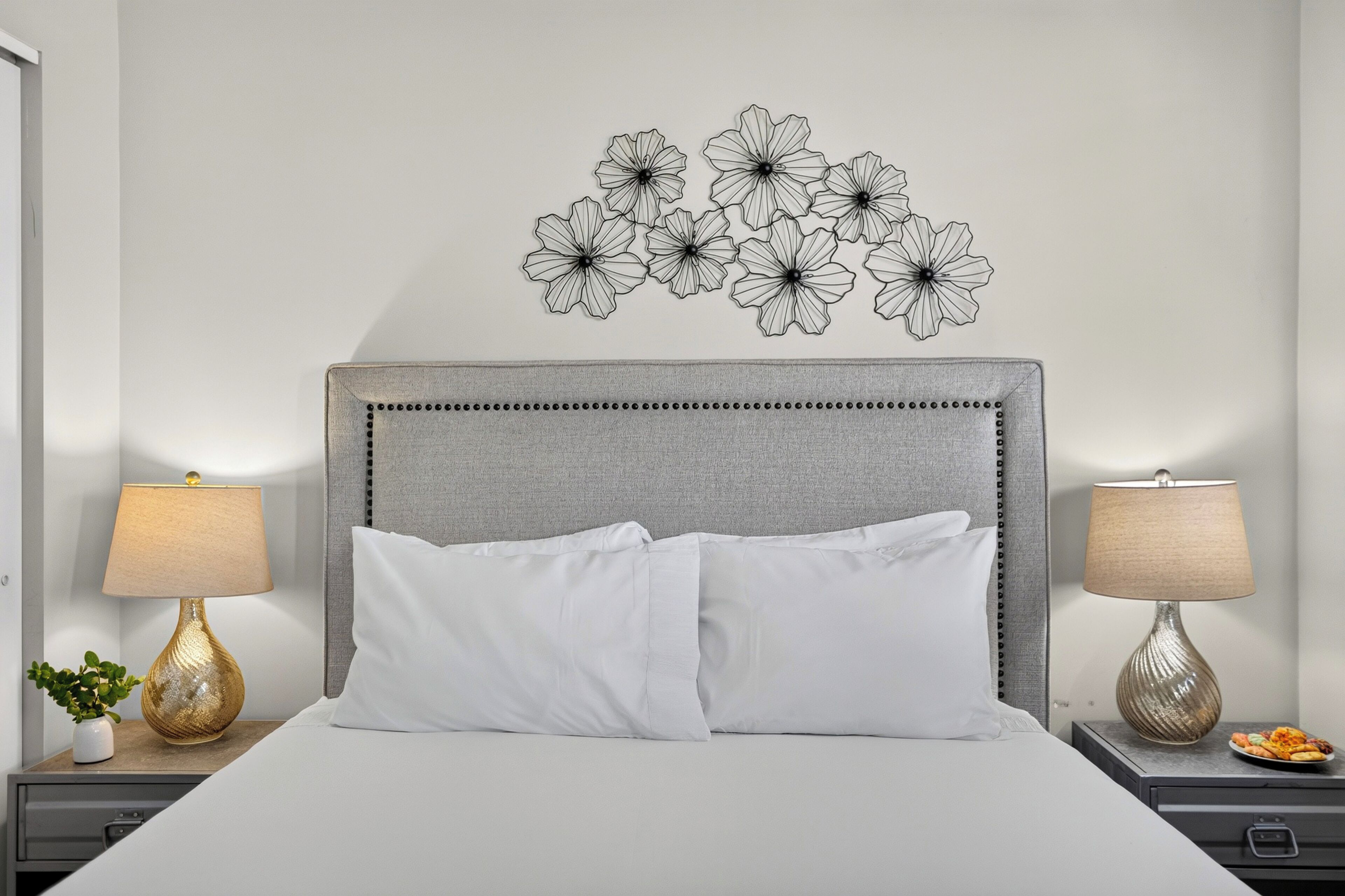 Un lit bien fait, avec du linge de lit blanc, deux lampes de chevet, un élément décoratif mural et une petite plante posée sur la table de chevet.