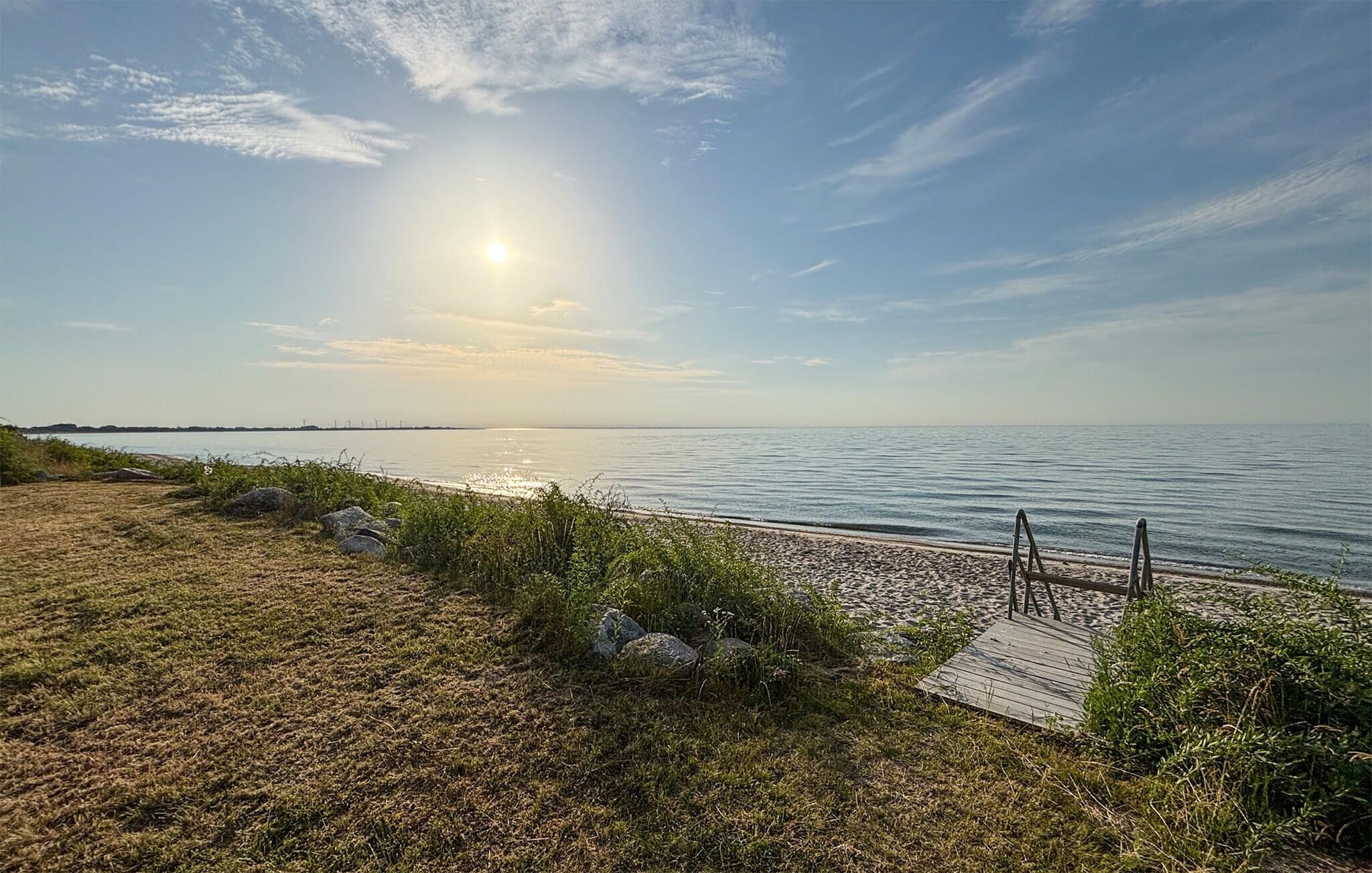 A beach with a wooden boardwalk leading to the water, a clear sky, and distant wind turbines.