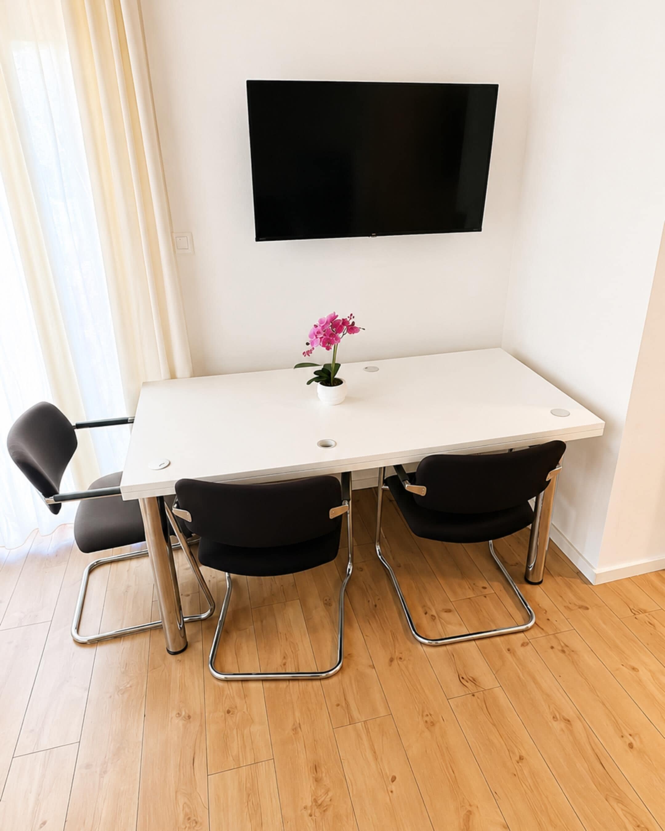 Een witte tafel met twee zwarte stoelen, een potplant en een flatscreen televisie aan de muur.