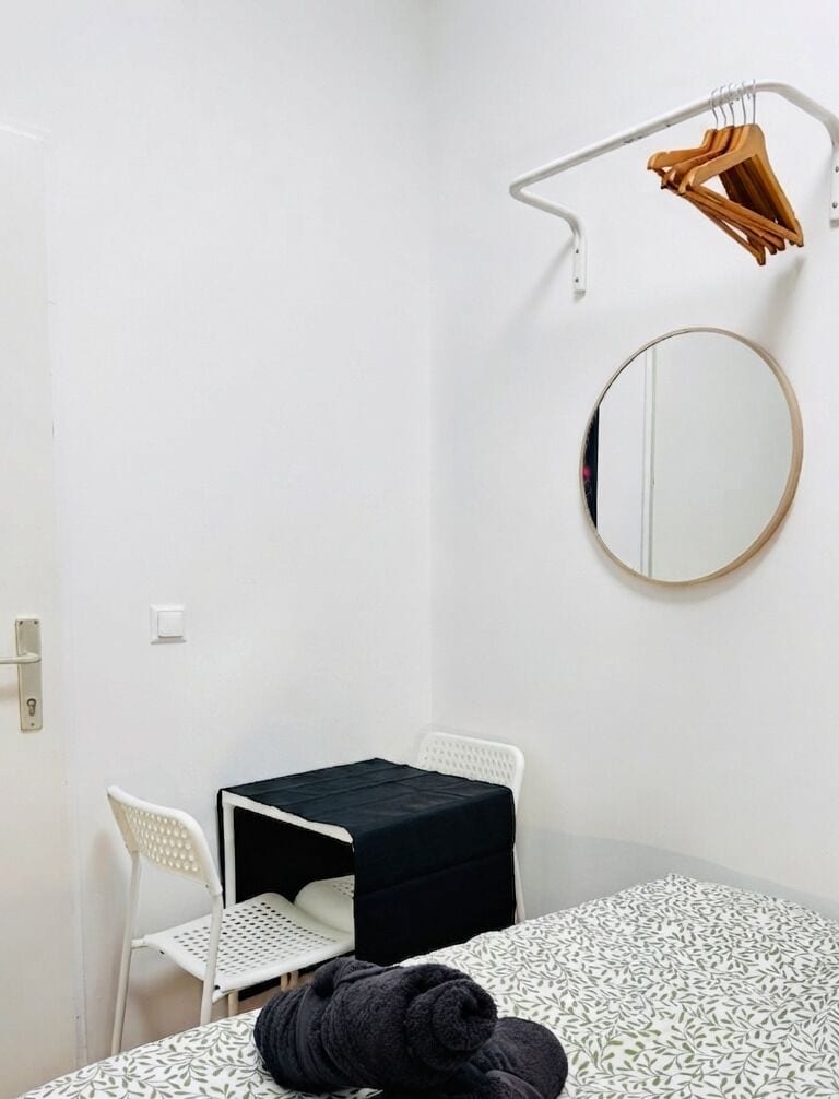 A small, white-walled room with a single bed, a round mirror, a small table, and a chair.