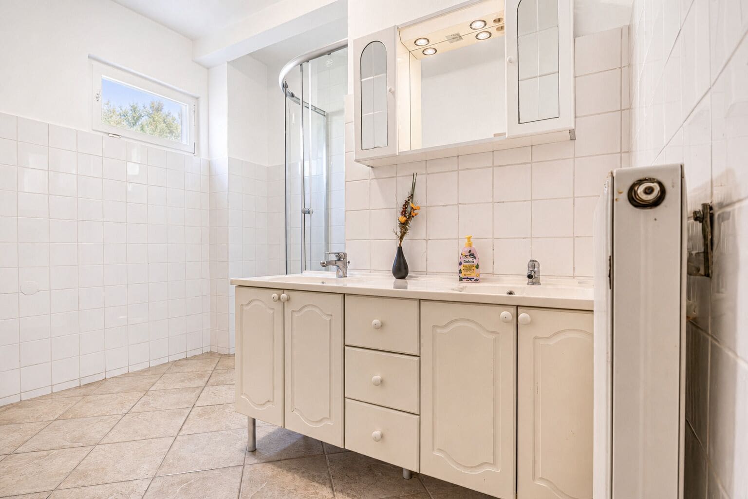 A bathroom with a white tiled floor, a beige vanity with a mirror, a shower, and a toilet.