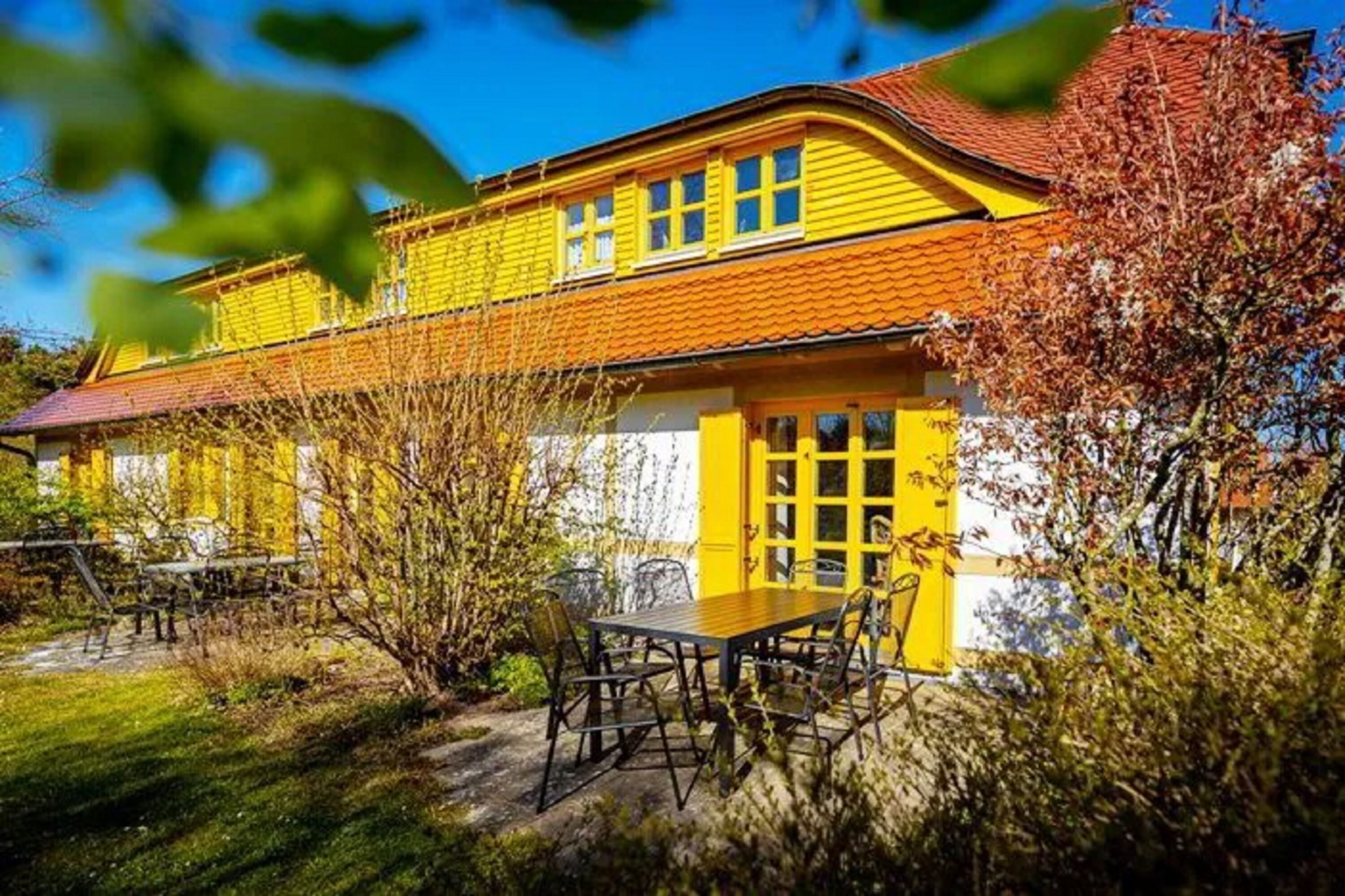 A yellow building with a red roof, a table with chairs, and a garden.
