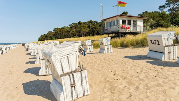 Een strand met rijen witte rieten strandstoelen en een bewakingspost met een vlag.