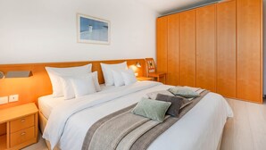 Een netjes opgemaakt bed met witte lakens en meerdere kussens, een houten nachtkastje met lades, een houten kledingkast en een ingelijst schilderij aan de muur.
