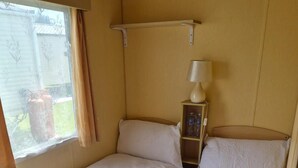 A room with two beds, a window with curtains, a bedside lamp, and a shelf with bottles. - Cabin Sleeps 6 With Access Outdoor Pool (Sandown)