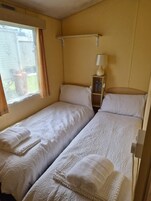 Una habitación con dos camas, una ventana con cortinas, una lámpara de noche y una estantería con botellas.