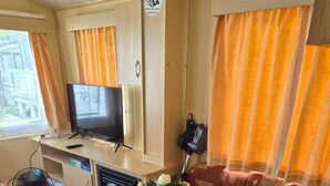 A cozy living room with a sofa, a television, a small table with a vase of flowers, and a fan. - Cabin Sleeps 6 With Access Outdoor Pool (Sandown)