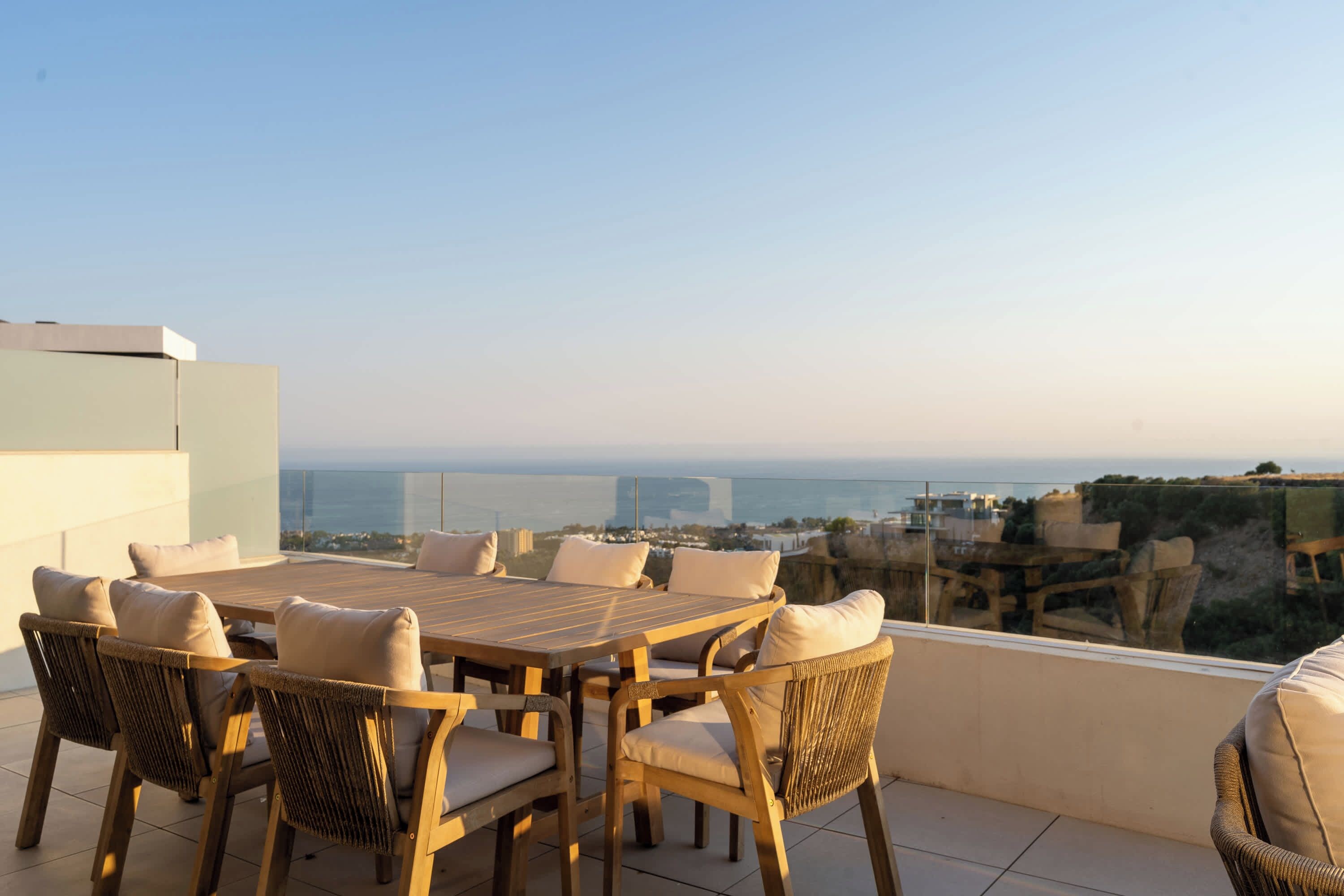 A terrace with a wooden table and wicker chairs, overlooking a coastal view.