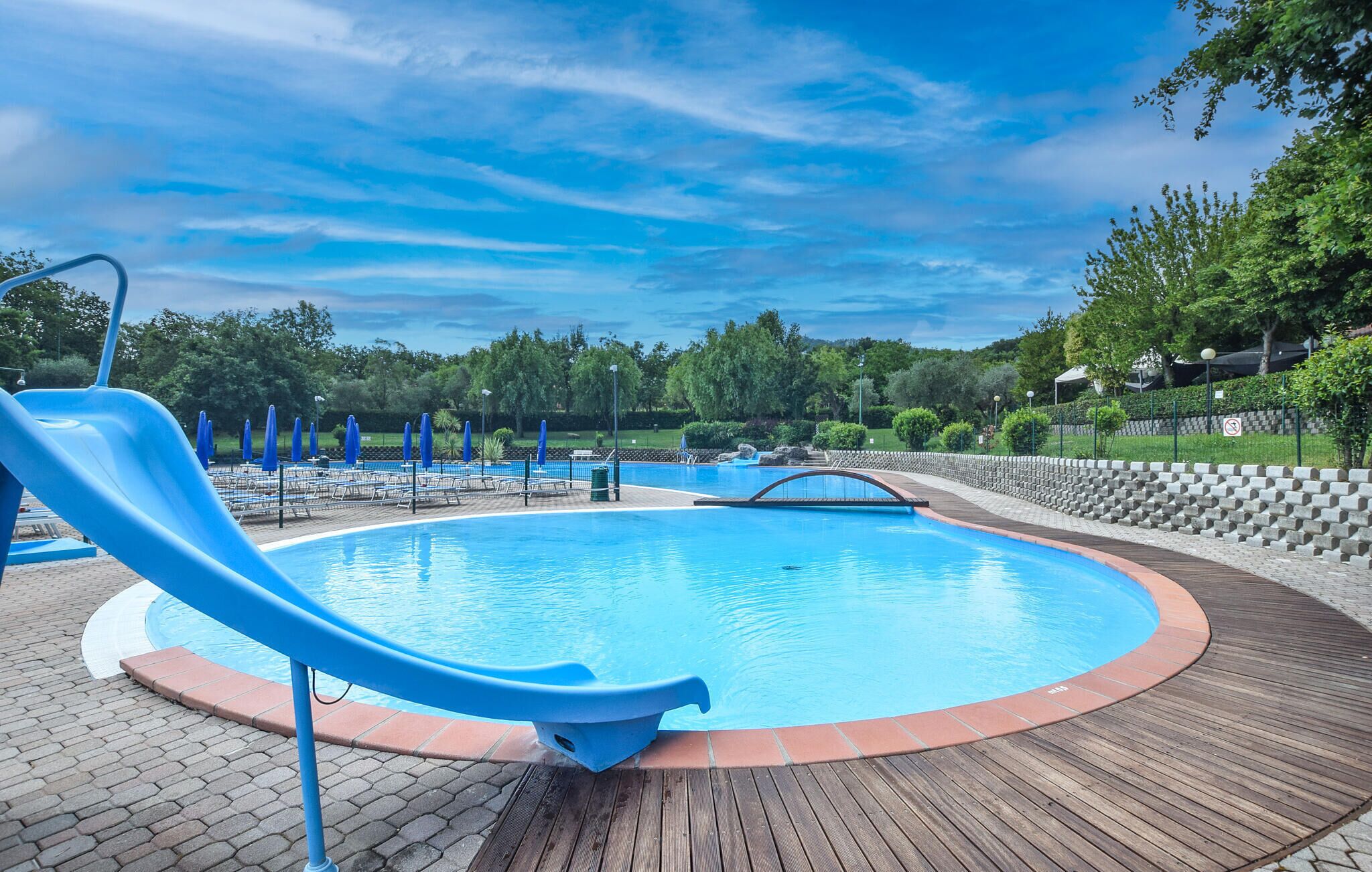 Piscina com escorregador, rodeada por um deck de madeira e um caminho de tijolos.