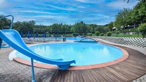 Uma piscina com escorrega, rodeada por um deck de tijolos e uma parede de pedra.